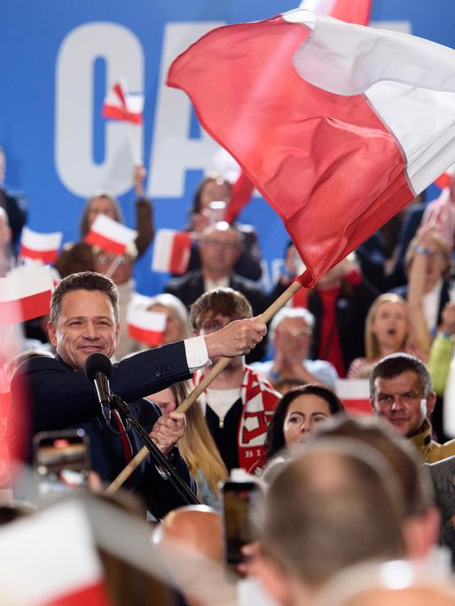 Rafał Trzaskowski schwenkt die polnische Flagge. Die Szene spielt sich am Sonntagabend, dem 18. Mai 2025, in Sandomierz ab. Im Hintergrund jubeln Unterstützer, viele halten kleine und große Polenfahnen in die Höhe. 