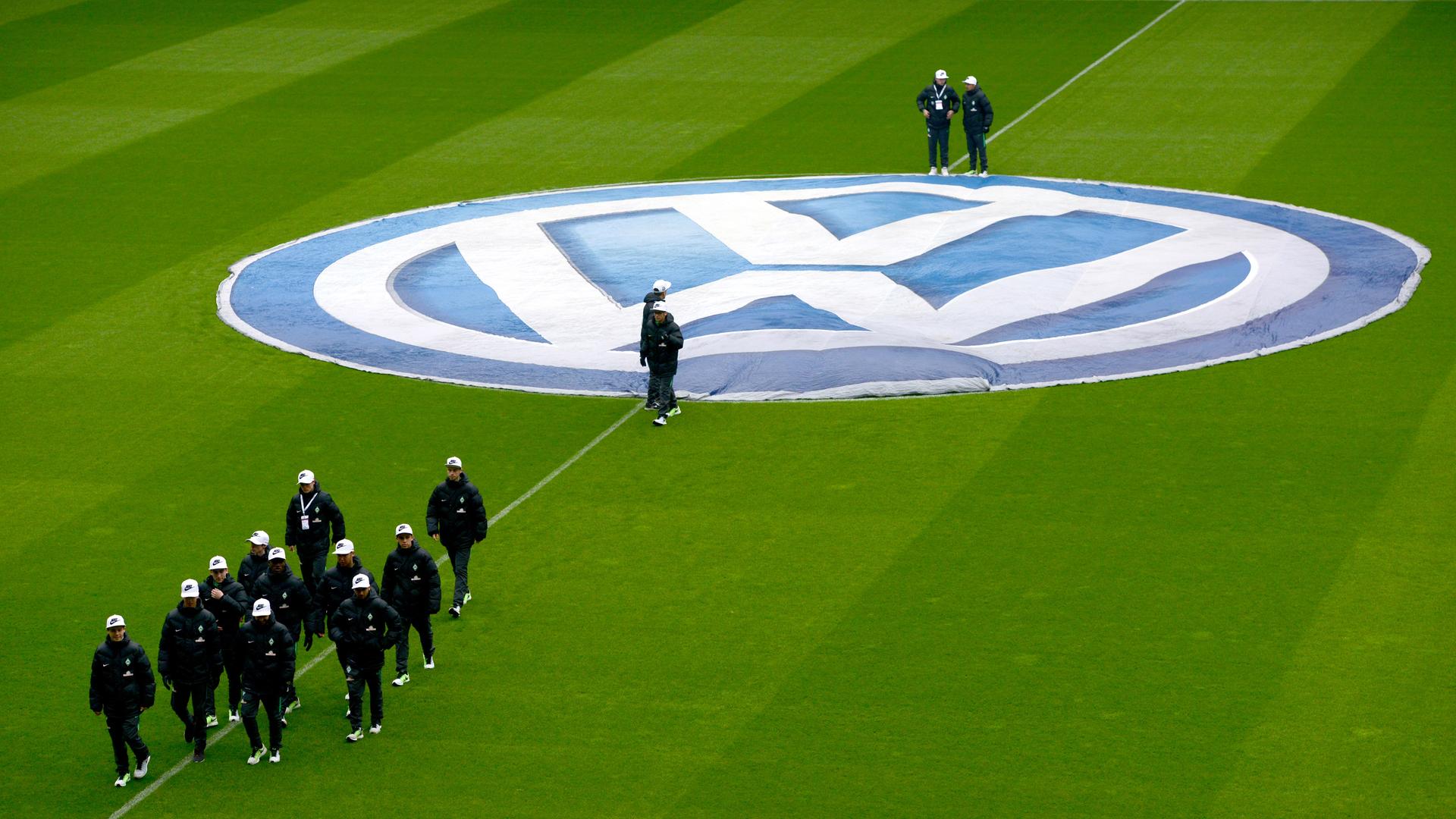 Blick von der Tribüne auf einen grünen Rasen mit dem VW-Logo über dem Anstoßkreis. Eine Gruppe von Balljungen mit weißen Baseballkappen läuft zur Außenlinie.