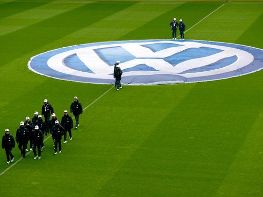 Blick von der Tribüne auf einen grünen Rasen mit dem VW-Logo über dem Anstoßkreis. Eine Gruppe von Balljungen mit weißen Baseballkappen läuft zur Außenlinie.