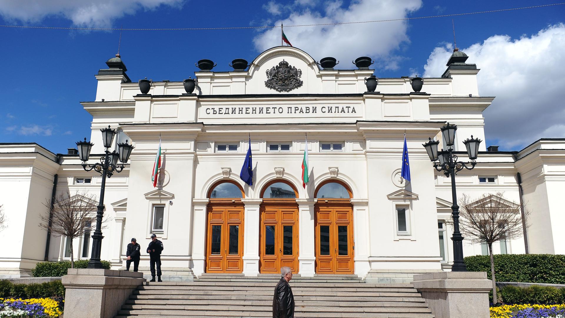 Das weiße Parlamentsgebäude hat drei braune Eingangstüren und ist mit Flaggen geschmückt. Mehrere Stufen führen zum Eingang.