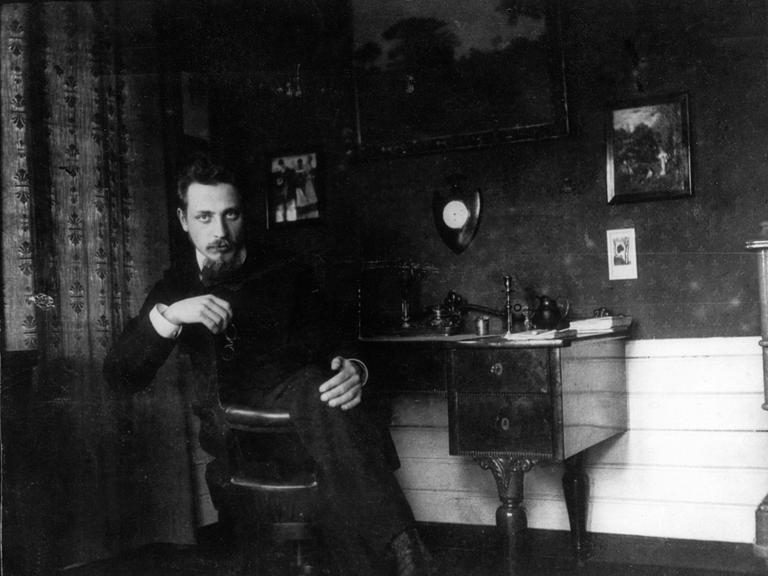 Der Dichter Rainer Maria Rilke sitzt um 1905 auf einem Stuhl in einem Zimmer.