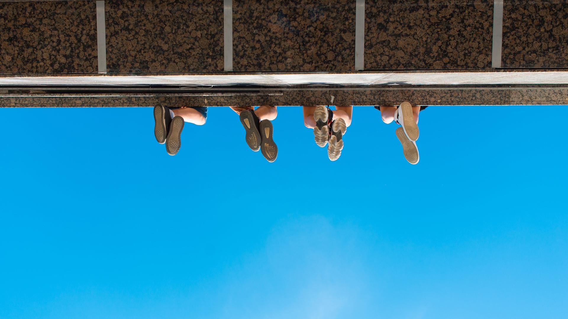 Unteransicht eines Absatzes wie bei einem Balkon oder einer Brücke. Zu sehen sind vier paar Füße von unten, wie sie von der Konstruktion herabbaumeln.