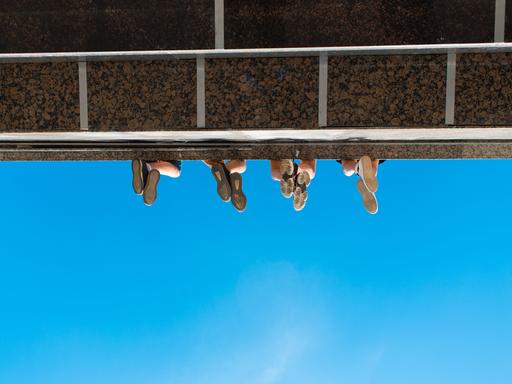 Unteransicht eines Absatzes wie bei einem Balkon oder einer Brücke. Zu sehen sind vier paar Füße von unten, wie sie von der Konstruktion herabbaumeln.