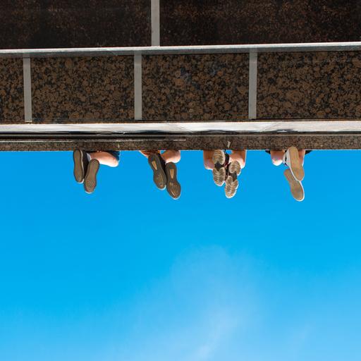Unteransicht eines Absatzes wie bei einem Balkon oder einer Brücke. Zu sehen sind vier paar Füße von unten, wie sie von der Konstruktion herabbaumeln.