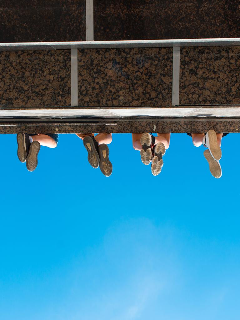 Unteransicht eines Absatzes wie bei einem Balkon oder einer Brücke. Zu sehen sind vier paar Füße von unten, wie sie von der Konstruktion herabbaumeln.