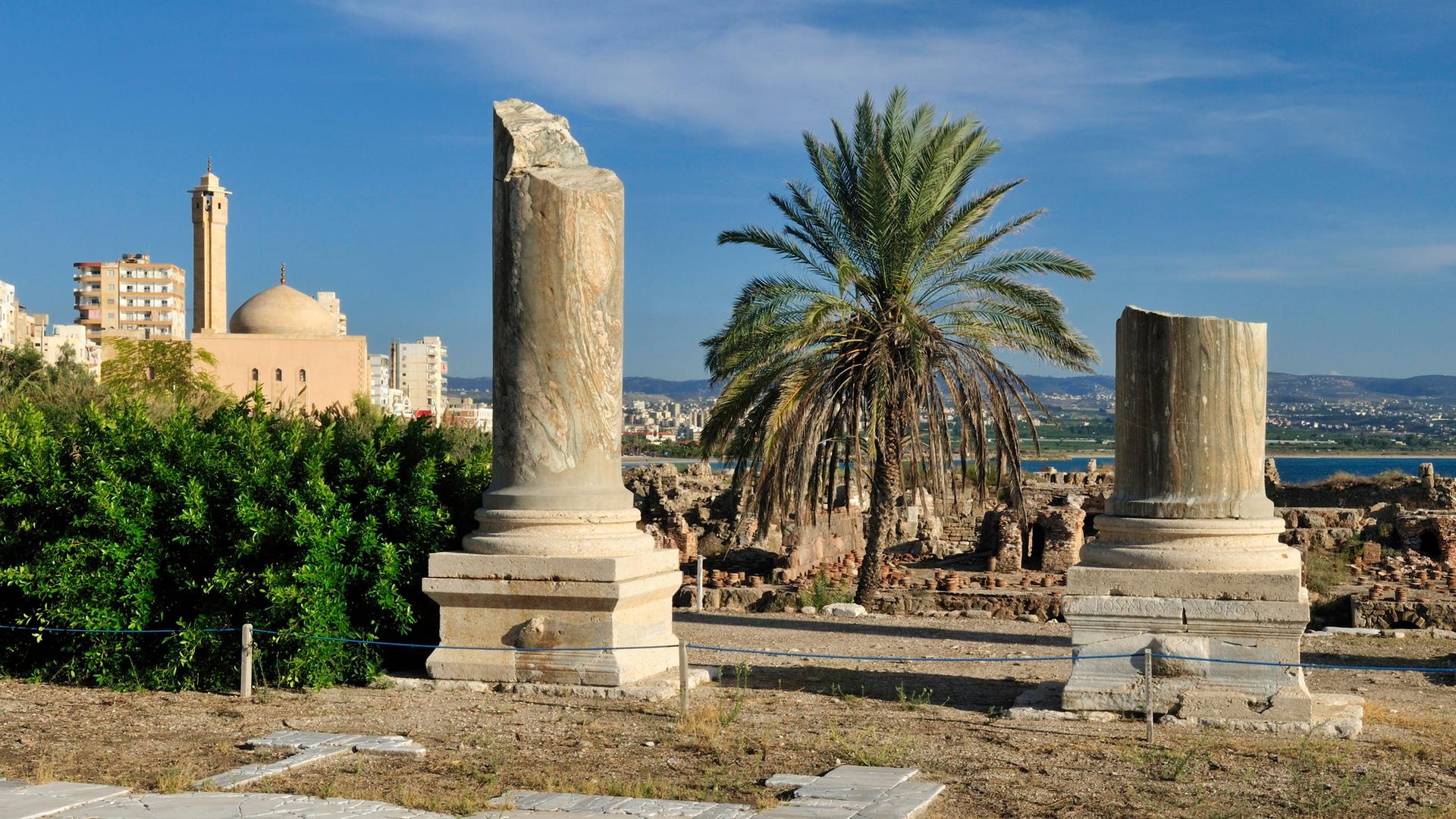 Archäologische Ausgrabungsstätte von Tyros im Libanon. Zwei große antike Säulen stehen zwischen Palmen und einer Moschee. Archäologische Ausgrabungsstätte von Tyros im Libanon. Zwei große antike Säulen stehen zwischen Palmen und einer Moschee.