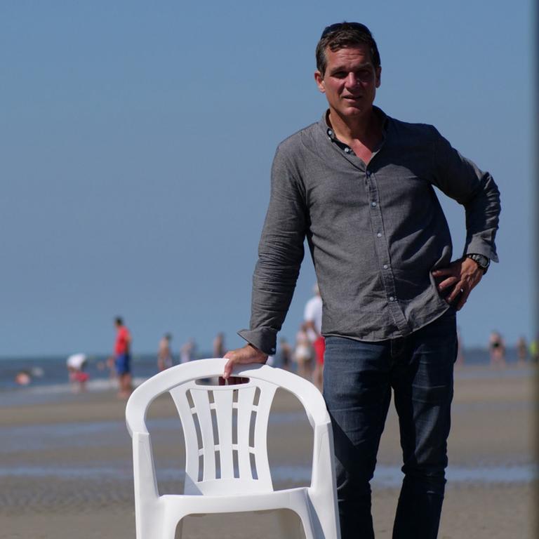 Der Autor Hauke Wendler steht an einem Sandstrand und stützt seinen rechten Arm auf einem weißen Plastikstuhl.