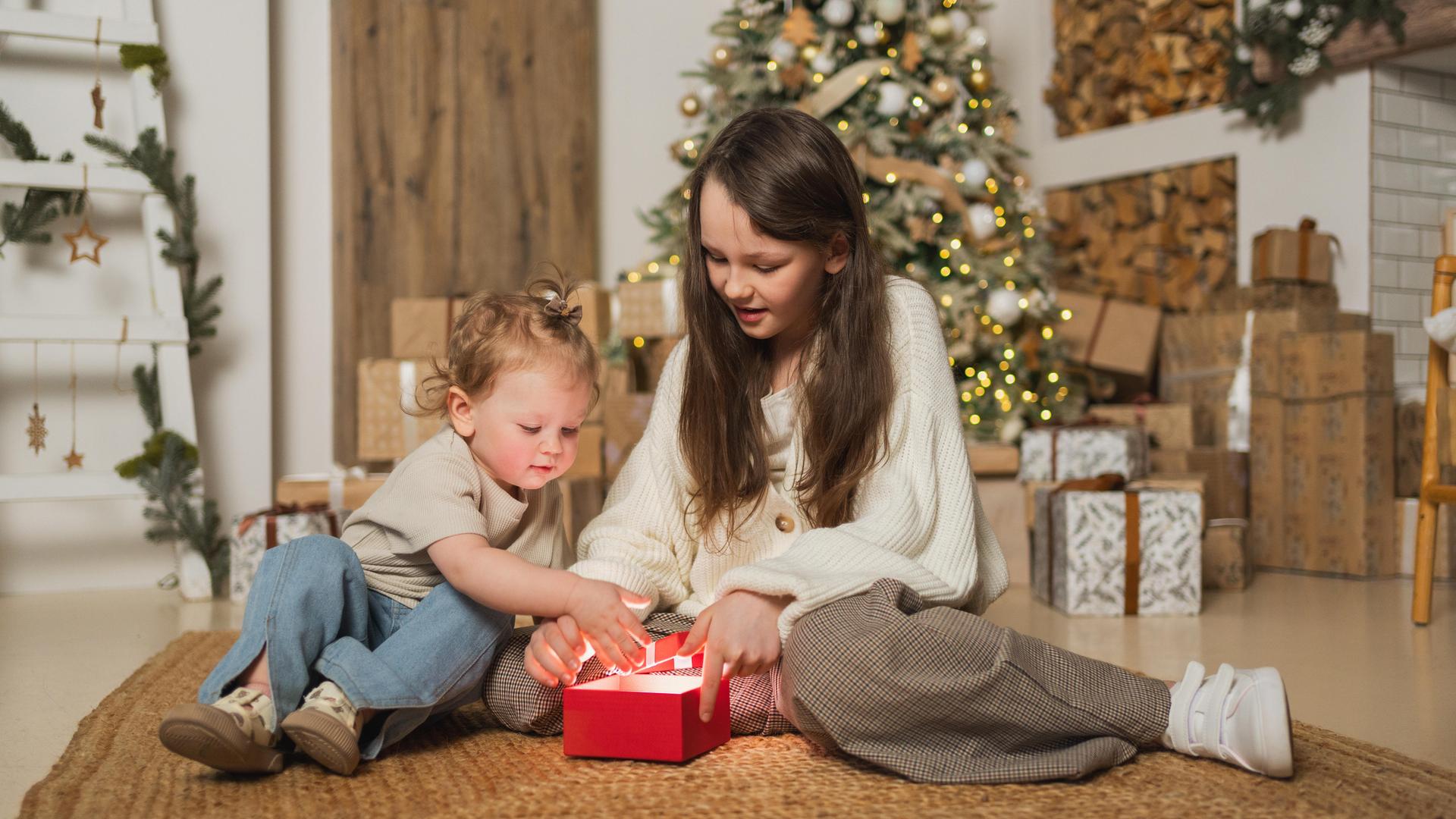 Ein großes und ein kleines Mädchen packen im Wohnzimmer zusammen ein Weihnachtsgeschenk aus. Ein großes und ein kleines Mädchen packen im Wohnzimmer zusammen ein Weihnachtsgeschenk aus.