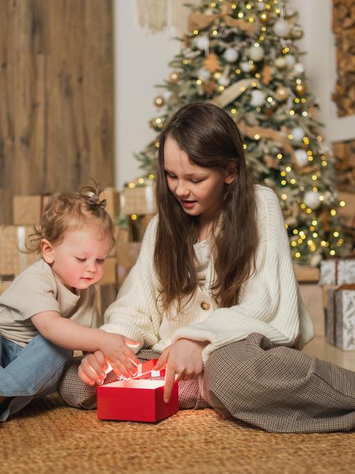 Ein großes und ein kleines Mädchen packen im Wohnzimmer zusammen ein Weihnachtsgeschenk aus. 