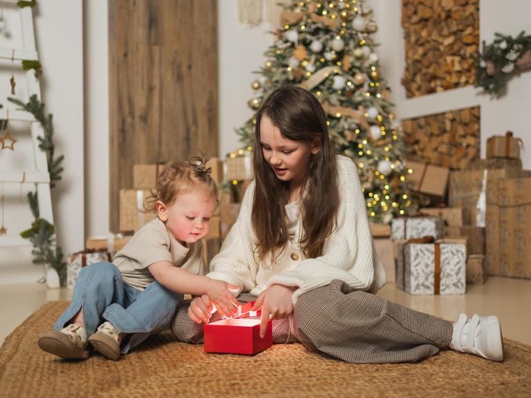 Ein großes und ein kleines Mädchen packen im Wohnzimmer zusammen ein Weihnachtsgeschenk aus. 