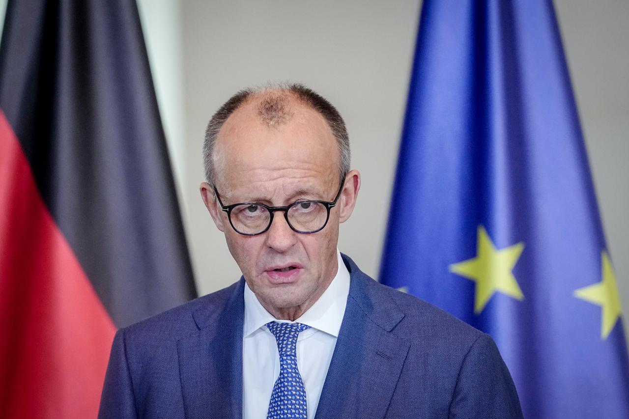 Das Bild zeigt Friedrich Merz mit einem ernsten Gesichtsausdruck vor einer deutschen und einer europäischen Flagge, während er ein Statement abgibt.