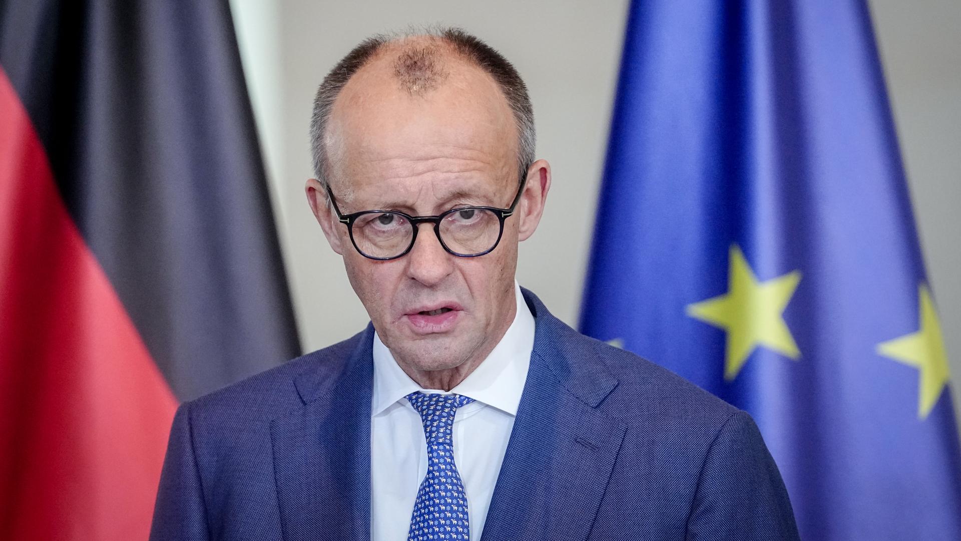 Das Bild zeigt Friedrich Merz mit einem ernsten Gesichtsausdruck vor einer deutschen und einer europäischen Flagge, während er ein Statement abgibt.