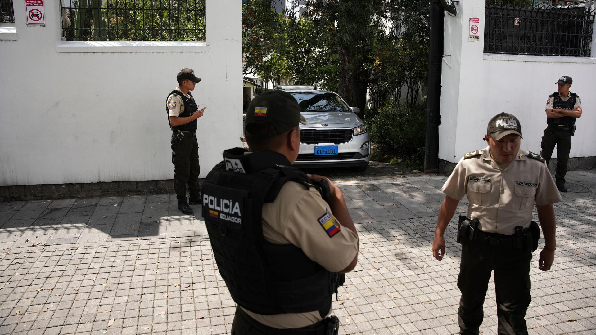 Vor der weißen Botschaftsmauer stehen vier Polizisten in beigen Uniformen. Ein weißes Auto verlässt gerade durch eine Ausfahrt das Botschaftsgelände.