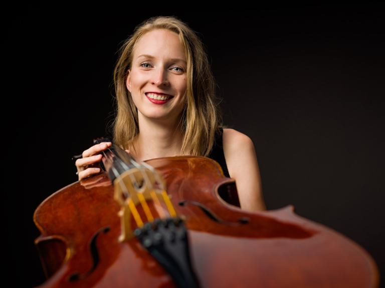 Marie-Elisaveth Hecker blickt lachend in die Kamera, während sie ihr Cello zur Kamera reckt. 