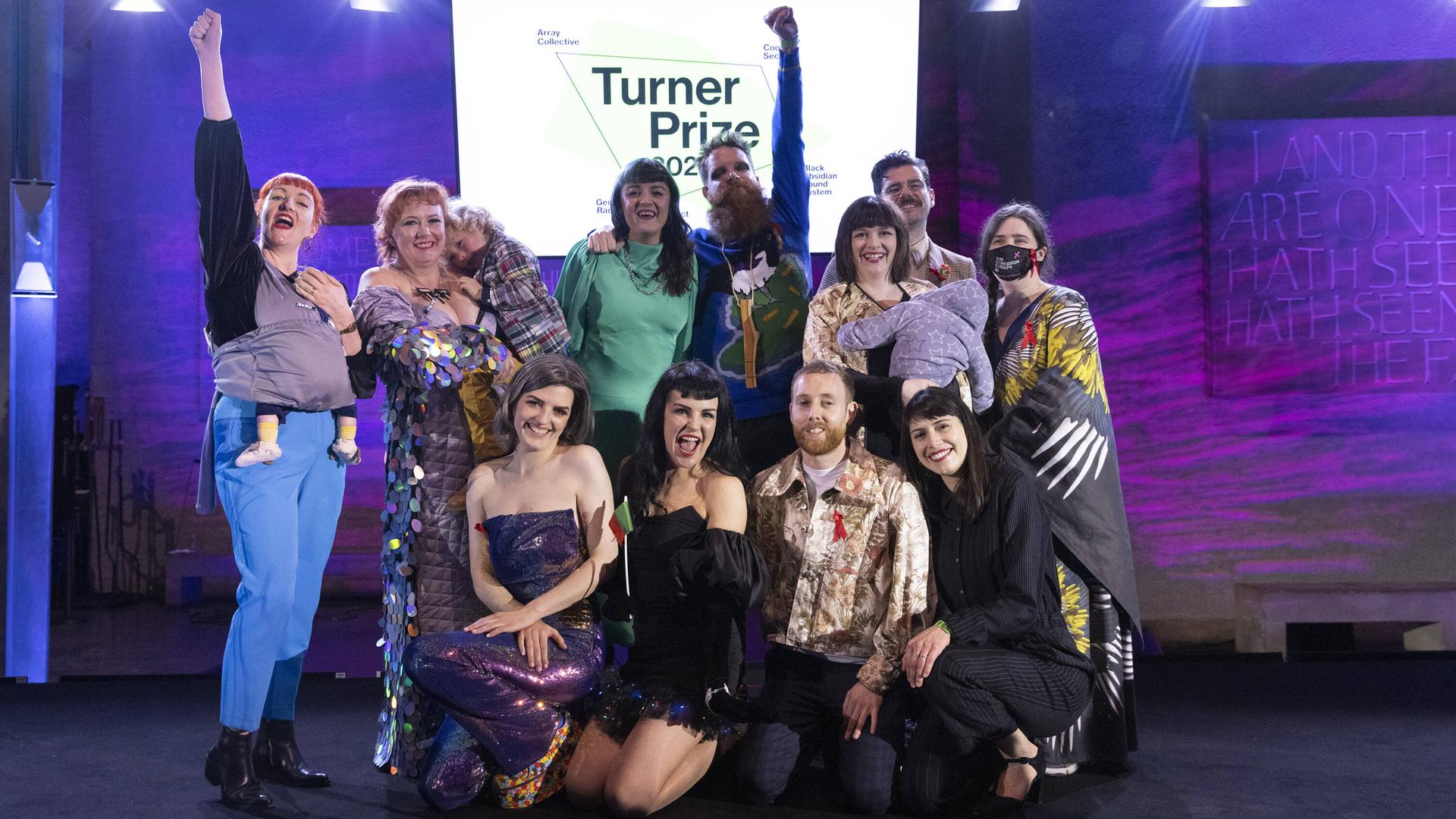 11 Menschen stehen auf einer Bühne vor einem Schild "Turner-Preis", einige recken die Faust in die Höhe, einige tragen Kinder auf dem Arm. Alle freuen sich. Es ist die Kunstgruppe Array Collective.