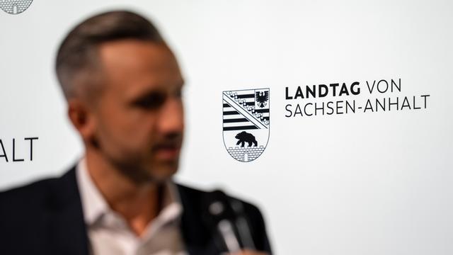 Ein Mann steht vor einer Wand mit der Aufschrift "Landtag von Sachsen-Anhalt". Er wird von einem Journalisten mit Mikrofon befragt, ist aber nur verschwommen zu erkennen. Es ist der AfD-Politiker Ulrich Siegmund.