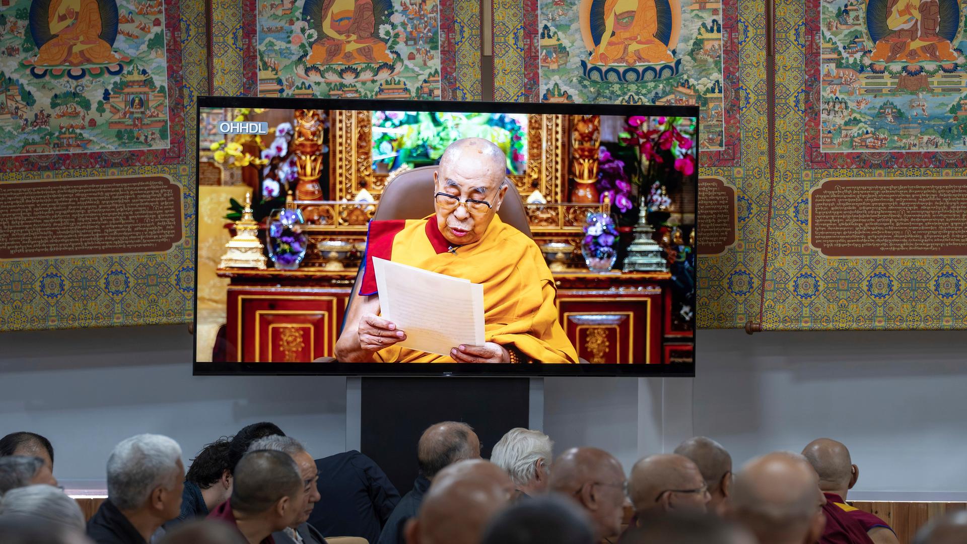 Buddhistische Mönche sitzen vor einem Fernseher und verfolgen eine Videobotschaft des Dalai Lama.