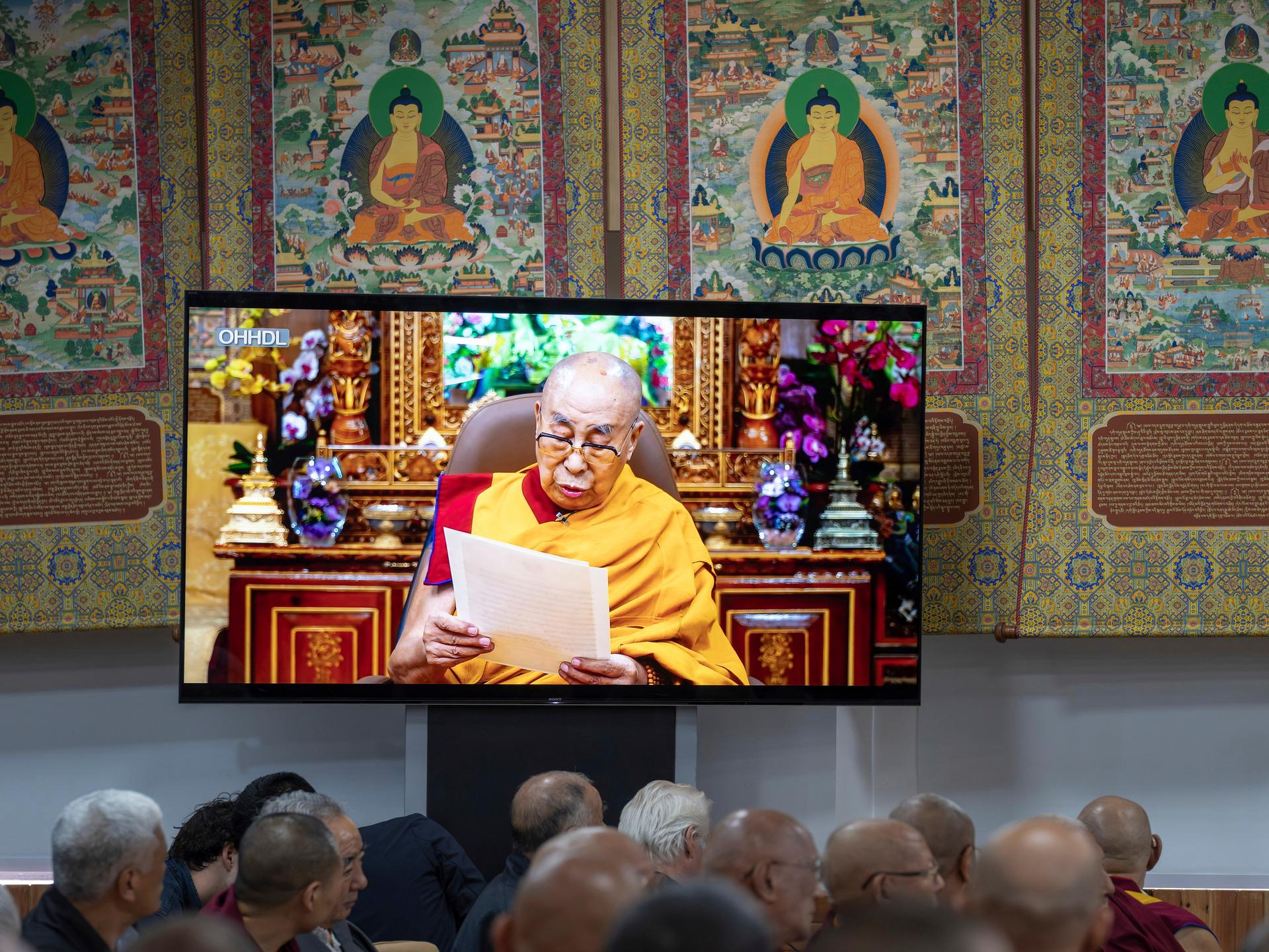 Buddhistische Mönche sitzen vor einem Fernseher und verfolgen eine Videobotschaft des Dalai Lama.
