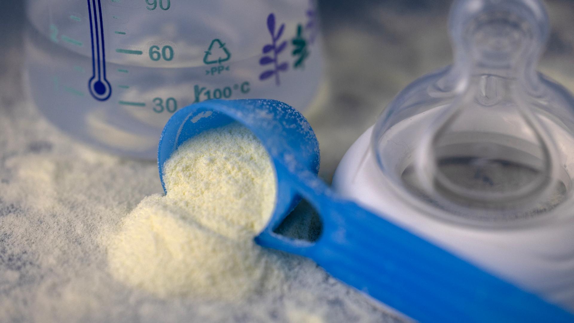 Man sieht einen blauen Dosierlöffel mit Babymilchpulver neben einer Babyflasche und einem Sauger. Das weiße Milchpulver liegt auch auf der Tischoberfläche.