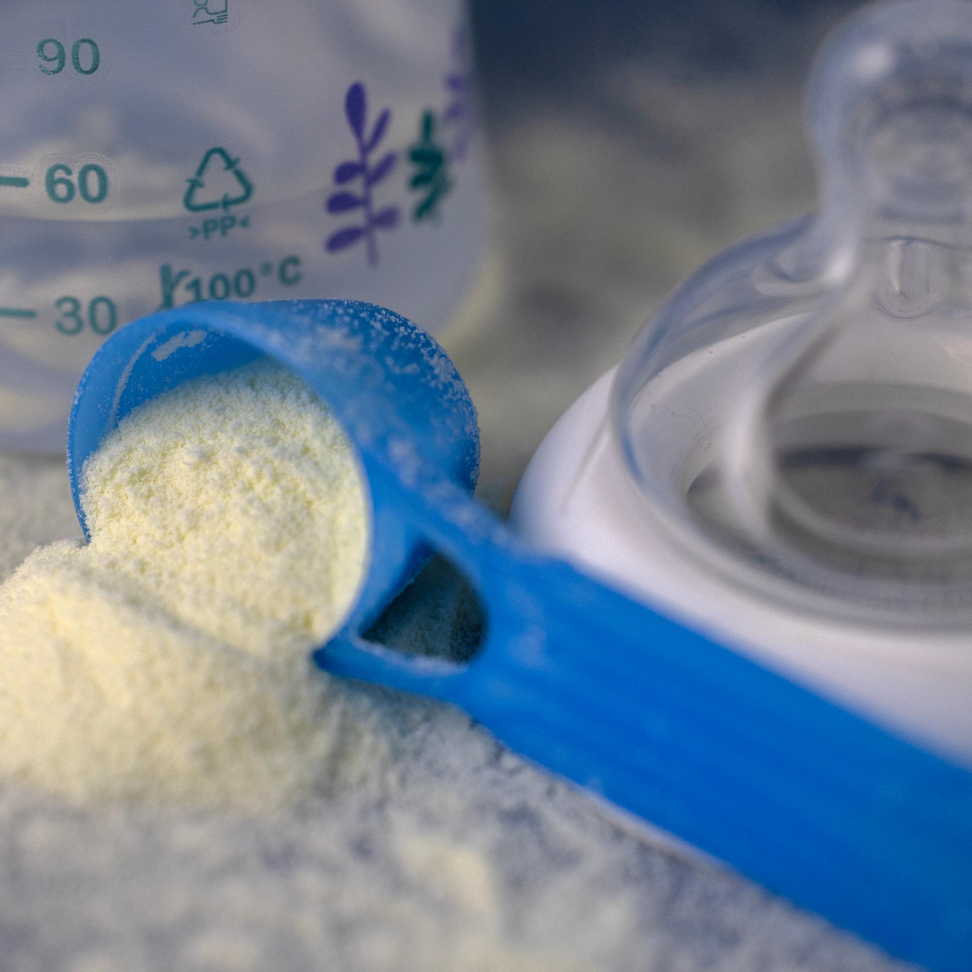 Man sieht einen blauen Dosierlöffel mit Babymilchpulver neben einer Babyflasche und einem Sauger. Das weiße Milchpulver liegt auch auf der Tischoberfläche.