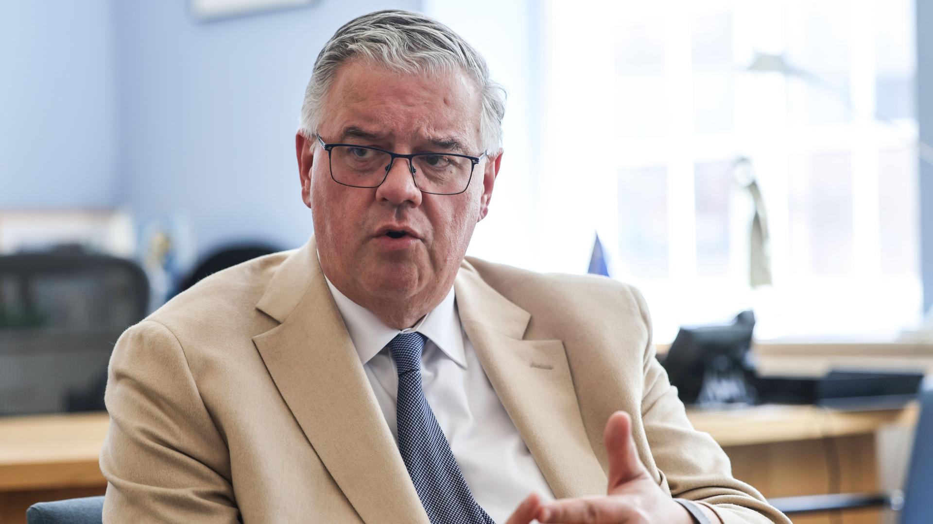 Hubert Bruls, Bürgermeister von Nimwegen und Chef der Euregio Rhein-Waal, blickt leicht an der Kamera vorbei während er spricht. Er trägt einen hellen Anzug, im Hintergrund ist ein Fenster. 