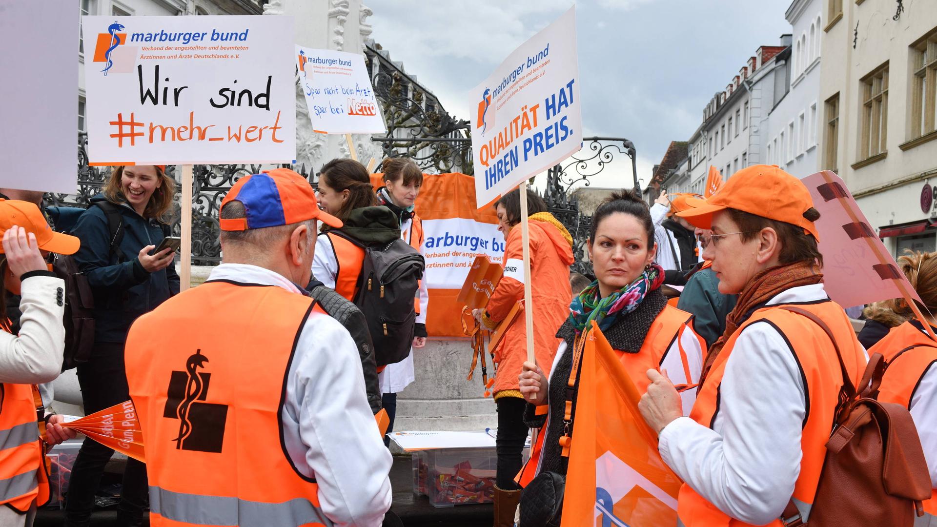Ärzte des Marburger Bundes demonstrieren für mehr Lohn. Sie tragen orange Warnwesten und Plakate mit Aufschriften wie: "Wir sind #mehr_wert".