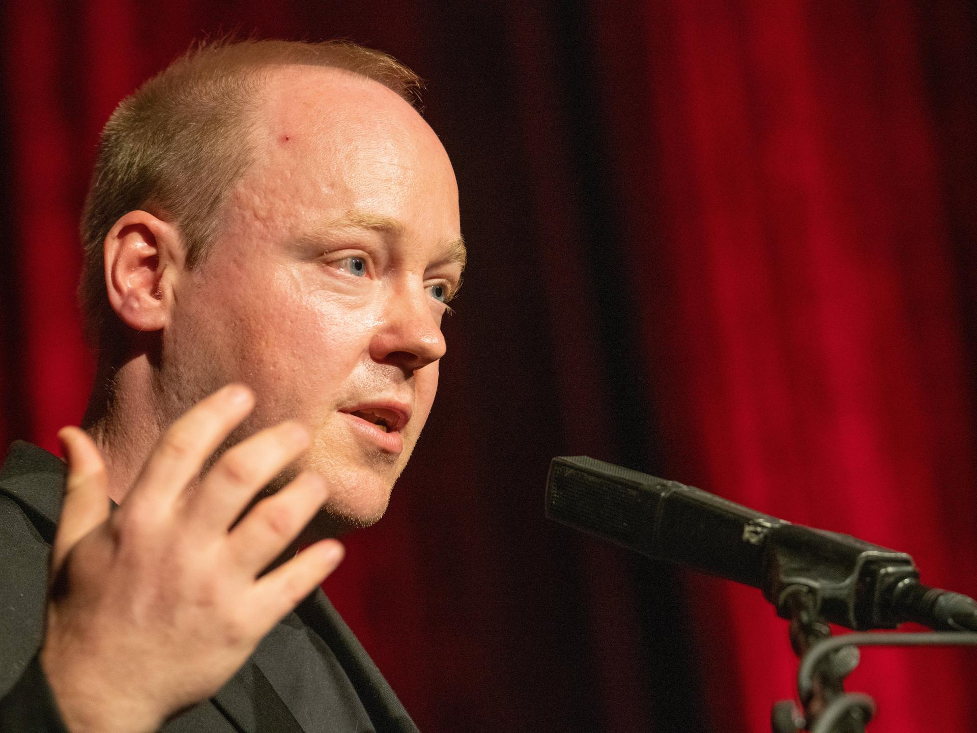 Christian Baron steht an einem Rednerpult und spricht gestikulierend in ein Mikrofon.
