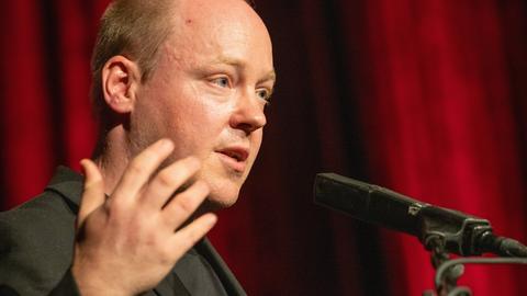Christian Baron steht an einem Rednerpult und spricht gestikulierend in ein Mikrofon.
