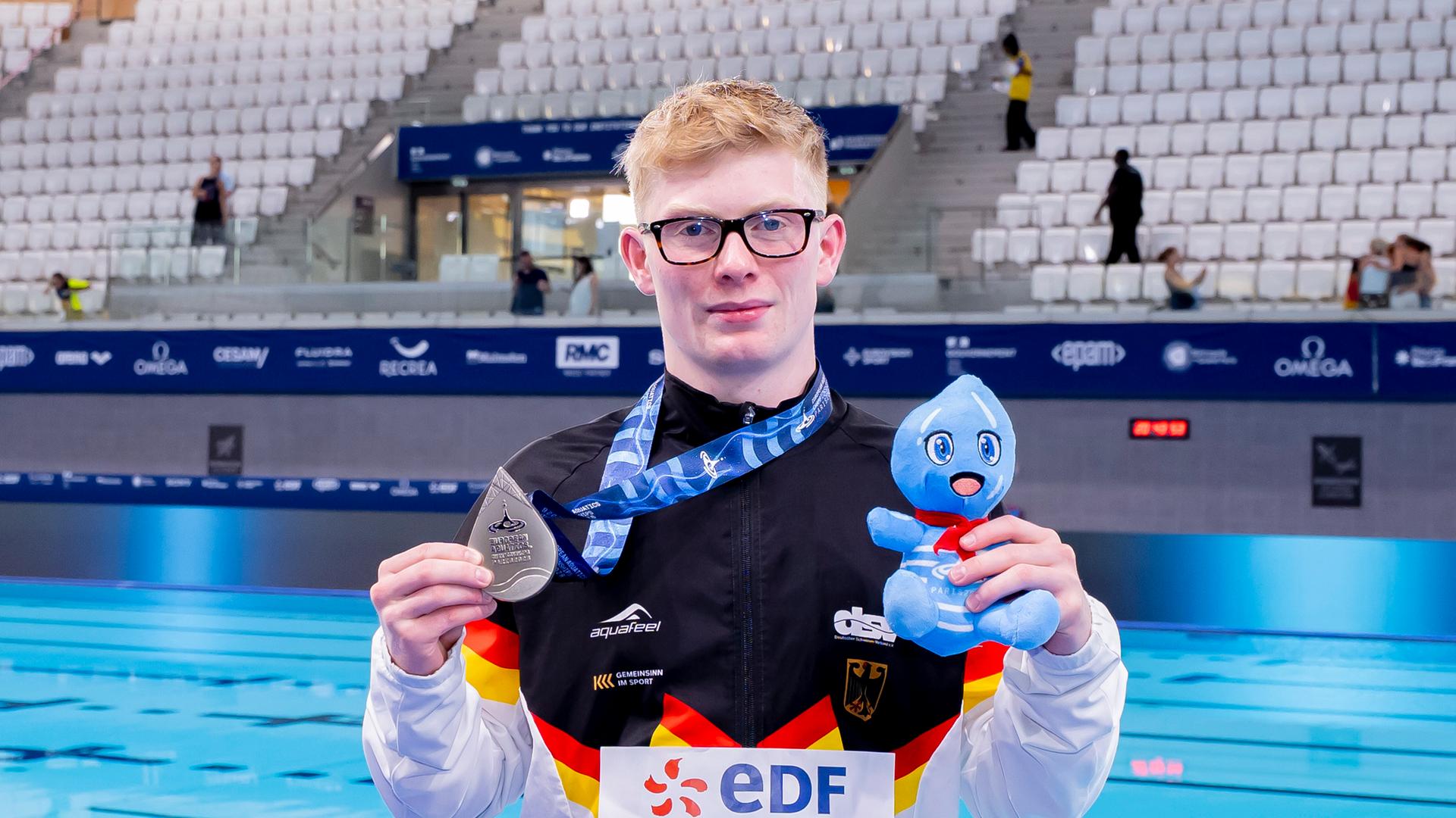 Jaden Eikermann steht am Beckenrand und zeigt seine Silbermedaille. 