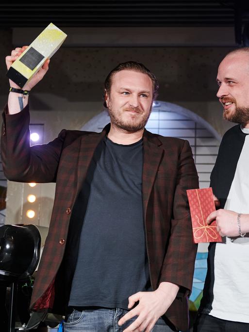 Hagen Decker und John Cook stehen auf einer Bühne und recken eine Trophäe in die Höhe.