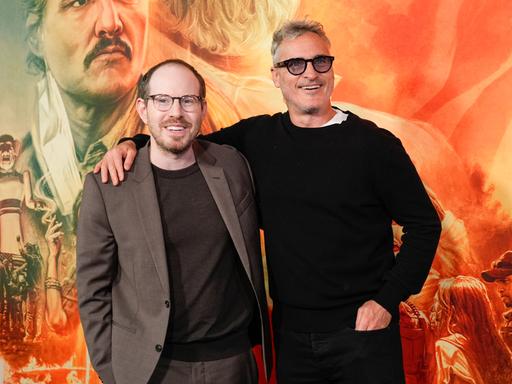 Regisseur Ari Aster (li.) und Schauspieler Joaquin Phoenix bei der Premiere ihres gemeinsamen Films Ellington. Beide lächeln vergnügt, im Hintergrund das überdimensionale Poster ihres Films. 