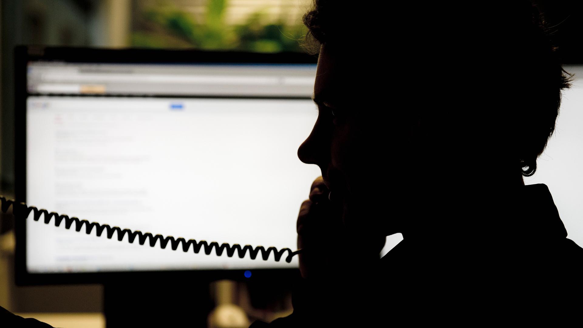 Silhouette eines Mannes im Profil, der vor einem Computerbildschirm sitzt und telefoniert.
