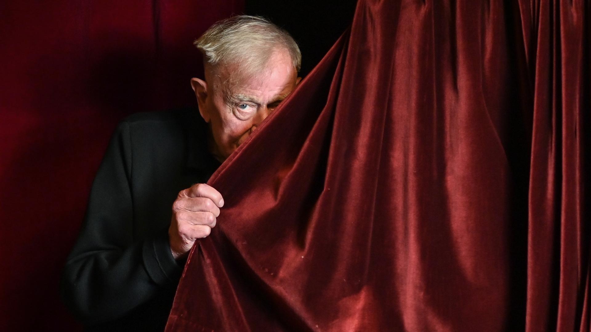 Der Regisseur Claus Peymann lugt hinter einem roten Samtvorhang hervor.