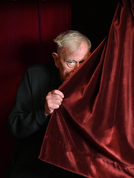 Der Regisseur Claus Peymann lugt hinter einem roten Samtvorhang hervor.