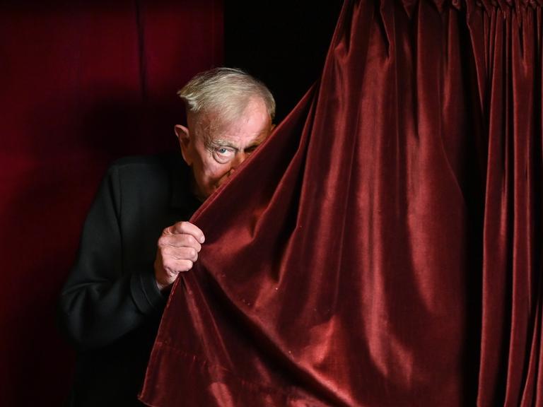 Der Regisseur Claus Peymann lugt hinter einem roten Samtvorhang hervor.