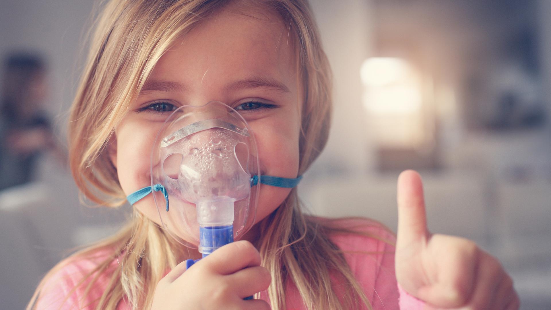 Ein Mädchen mit Atemmaske für Asthma lächelt in die Kamera und hält den Daumen hoch Ein Mädchen mit Atemmaske für Asthma lächelt in die Kamera und hält den Daumen hoch
