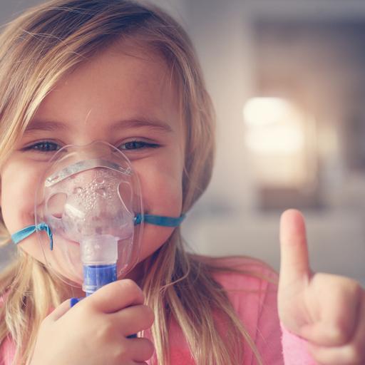 Ein Mädchen mit Atemmaske für Asthma lächelt in die Kamera und hält den Daumen hoch