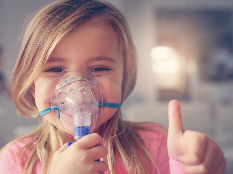 Ein Mädchen mit Atemmaske für Asthma lächelt in die Kamera und hält den Daumen hoch
