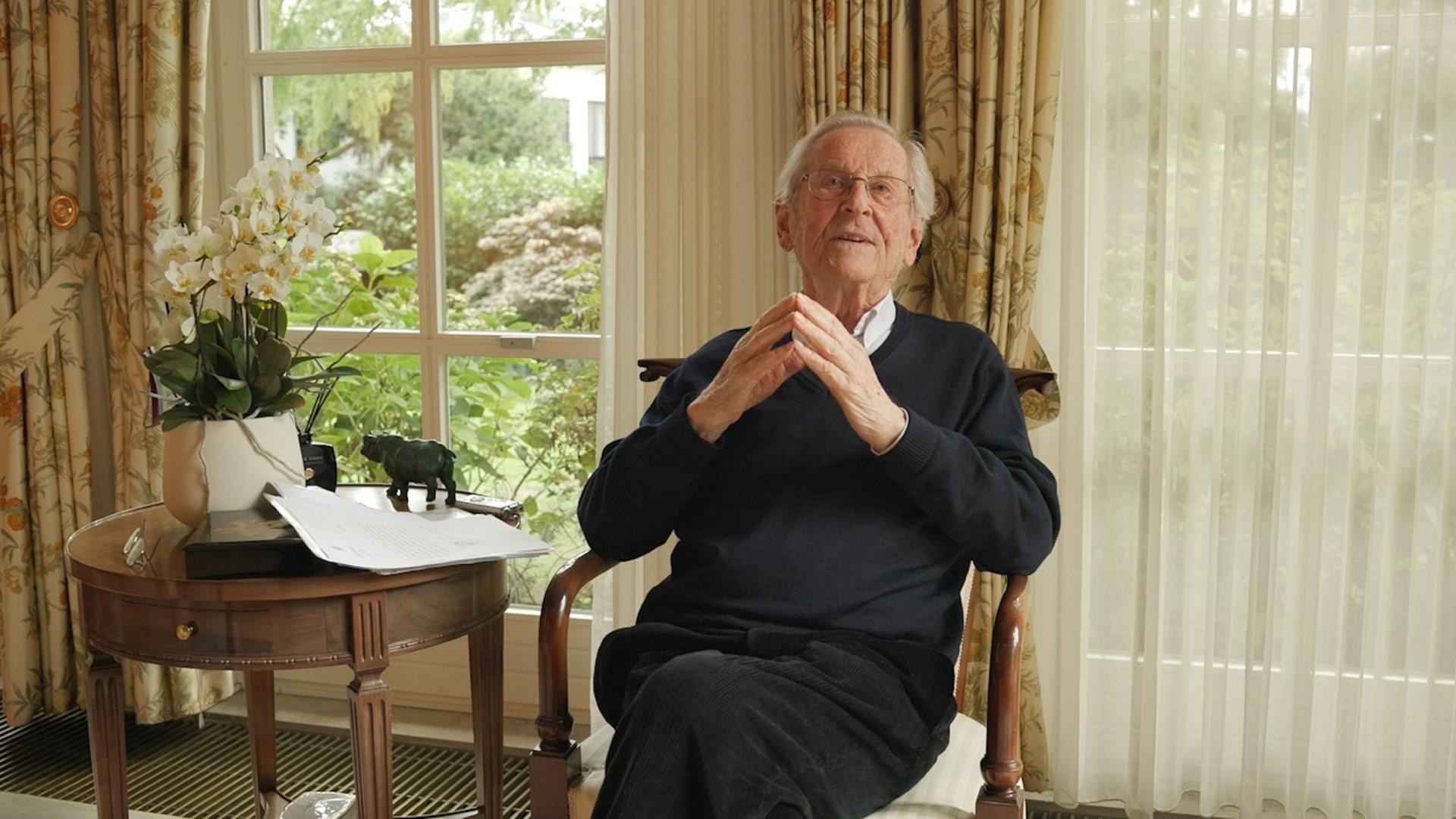 Klaus Adlung sitzt als alter Mann in einem Sessel in seinem Wohnzimmer, im Hintergrund ein Fenster mit Gardine. 