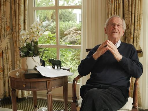 Klaus Adlung sitzt als alter Mann in einem Sessel in seinem Wohnzimmer, im Hintergrund ein Fenster mit Gardine. 