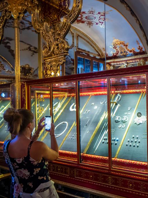 In einer Vitrine liegen aufgebahrte Juwelen. Eine Frau steht vor der Vitrine und fotografiert sie. Der Raum ist kunstvoll verziert.