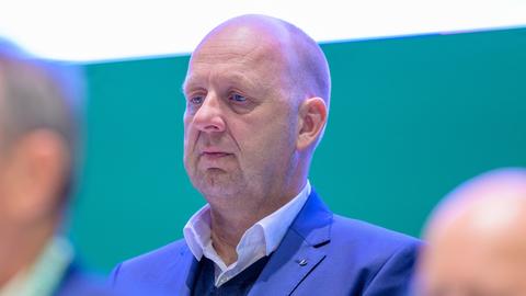 Ein Mann, Oliver Rau, mit raspelkurzen Haaren im Anzug im Halbprofil. Er trägt einen blauen Anzug und ein weißes Hemd ohne Krawatte.