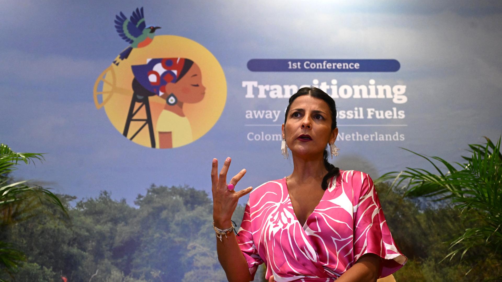 Ministerin Vélez Torres spricht auf einer Veranstaltung der Konferenz. Sie gestikuliert mit einer Hand und schaut ernst an der Kamera vorbei. Im Hintergrund ist ein Plakat mit dem Logo der Konferenz zu sehen. 