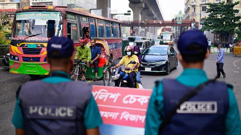 Sicherheitskräfte stehen an einer Verkehrskreuzung in Dhaka, Bangladesch. Zwei Mopeds, ein Auto und ein Bus stehen vor den Polizisten. 