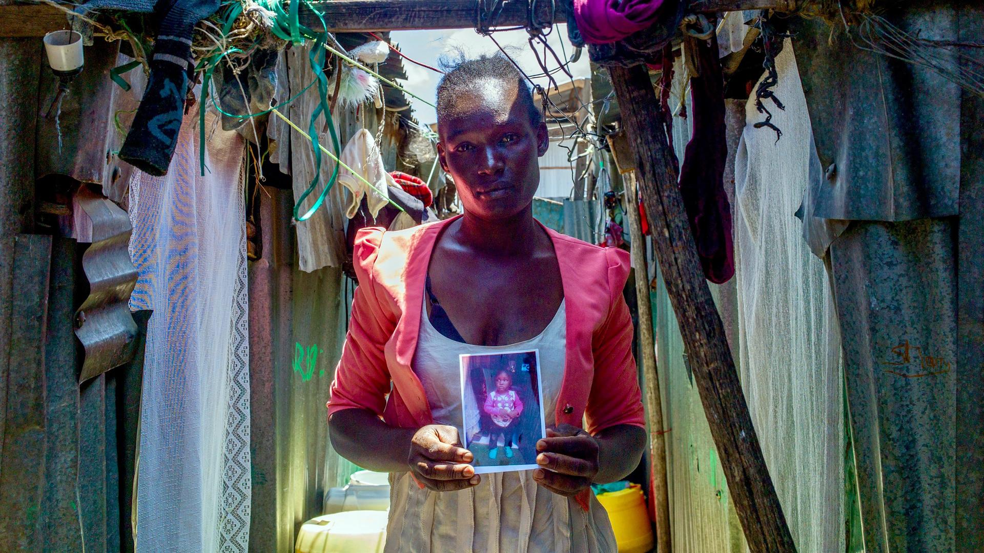 Die Mutter Lumbasi hält ein Foto ihres dreijährigen Sohnes in die Kamera und steht vor einer Hütte in einem Slum in Kenia.