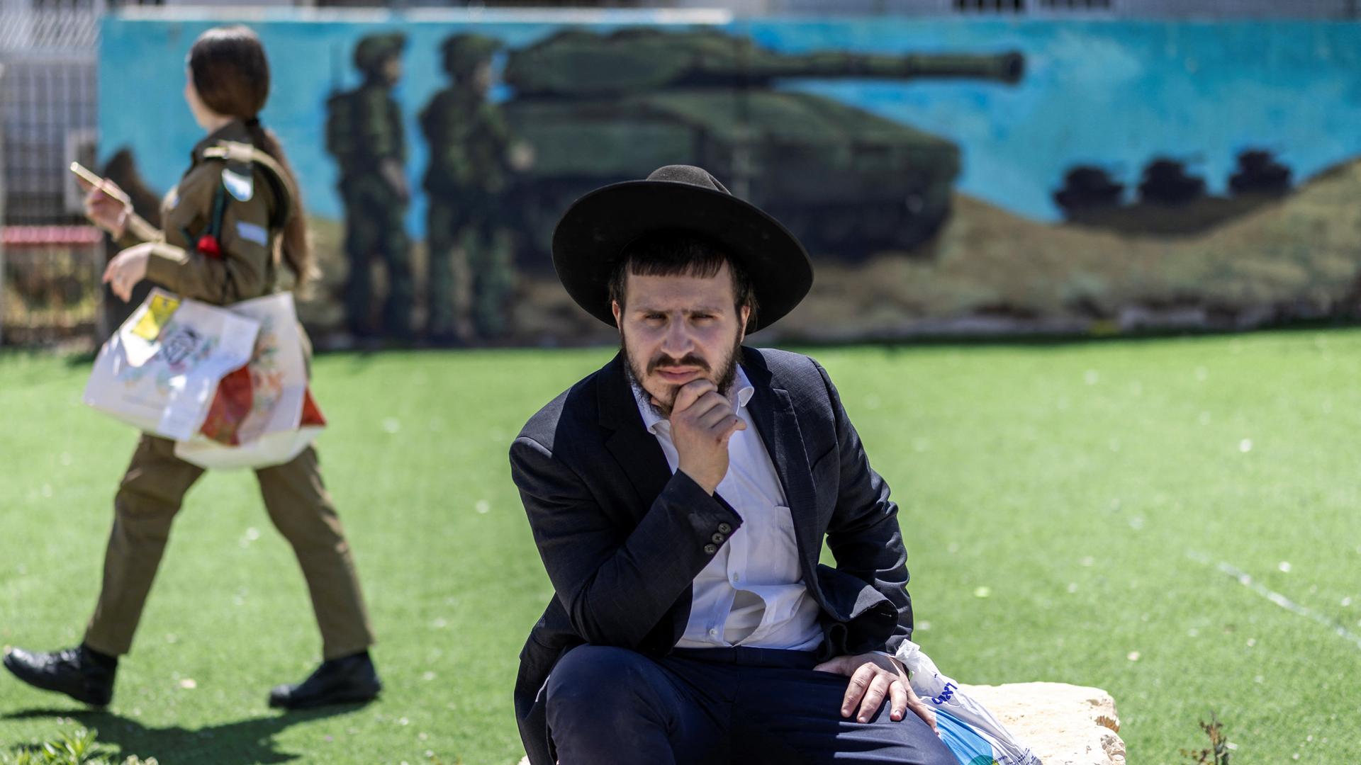 Eine israelische Soldatin läuft vor einem Wandgemälde, auf dem ein Panzer und Soldaten zu sehen sind. Im Vordergrund sitzt ein ultraorthodoxer Mann auf einem Stein. Eine israelische Soldatin läuft vor einem Wandgemälde, auf dem ein Panzer und Soldaten zu sehen sind. Im Vordergrund sitzt ein ultraorthodoxer Mann auf einem Stein.
