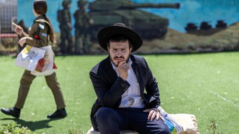 Eine israelische Soldatin läuft vor einem Wandgemälde, auf dem ein Panzer und Soldaten zu sehen sind. Im Vordergrund sitzt ein ultraorthodoxer Mann auf einem Stein. Eine israelische Soldatin läuft vor einem Wandgemälde, auf dem ein Panzer und Soldaten zu sehen sind. Im Vordergrund sitzt ein ultraorthodoxer Mann auf einem Stein.