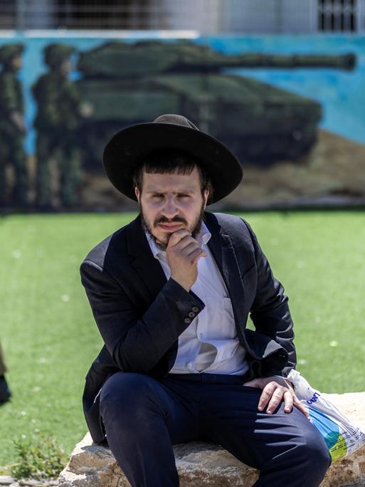 Eine israelische Soldatin läuft vor einem Wandgemälde, auf dem ein Panzer und Soldaten zu sehen sind. Im Vordergrund sitzt ein ultraorthodoxer Mann auf einem Stein.