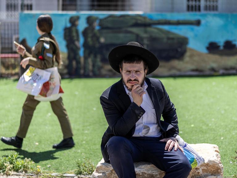 Eine israelische Soldatin läuft vor einem Wandgemälde, auf dem ein Panzer und Soldaten zu sehen sind. Im Vordergrund sitzt ein ultraorthodoxer Mann auf einem Stein.
