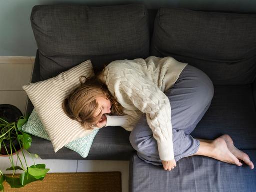 Eine blonde junge Frau liegt zusammengekauert auf einem Sofa. Ihre Beine sind angewinkelt und sie umfasst ihre Knie mit ihren Armen.